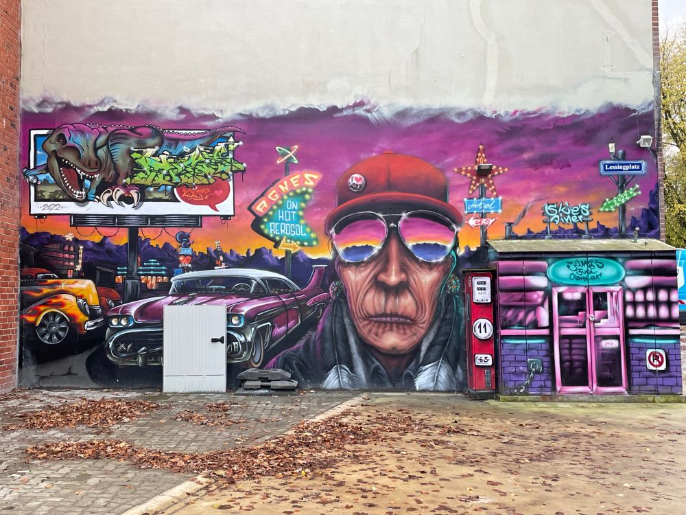 Eine Häuserwand mit Street Art im amerikanischen Stil. Zu sehen sind unter anderem der riesige Kopf einer anscheinend älteren Person mit Sonnenbrille und Cappy, fancy Autos, Leuchtreklame-Schilder und sogar ein T-Rex.