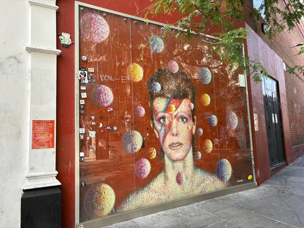 David Bowie Mural in Brixton, London, durch eine Glasfront geschützt. Es zeigt Bowies Kopf im Stil seines "Aladdin Sane" Albumcovers mit rot-blauem Blitz im Gesicht, um ihn herum viele bunte Kugeln.