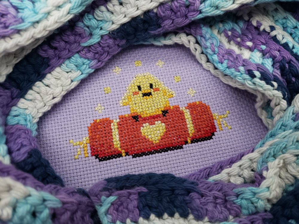 A cross stitch of a small chick sitting on an ornate red cushion.