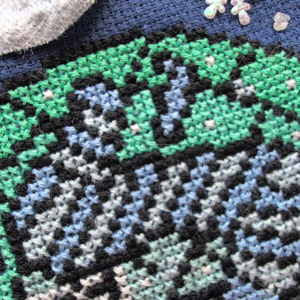 An extreme close up of a cross stitch on navy aida showing stitching of billowing white and blue steam against mint green stitches.
