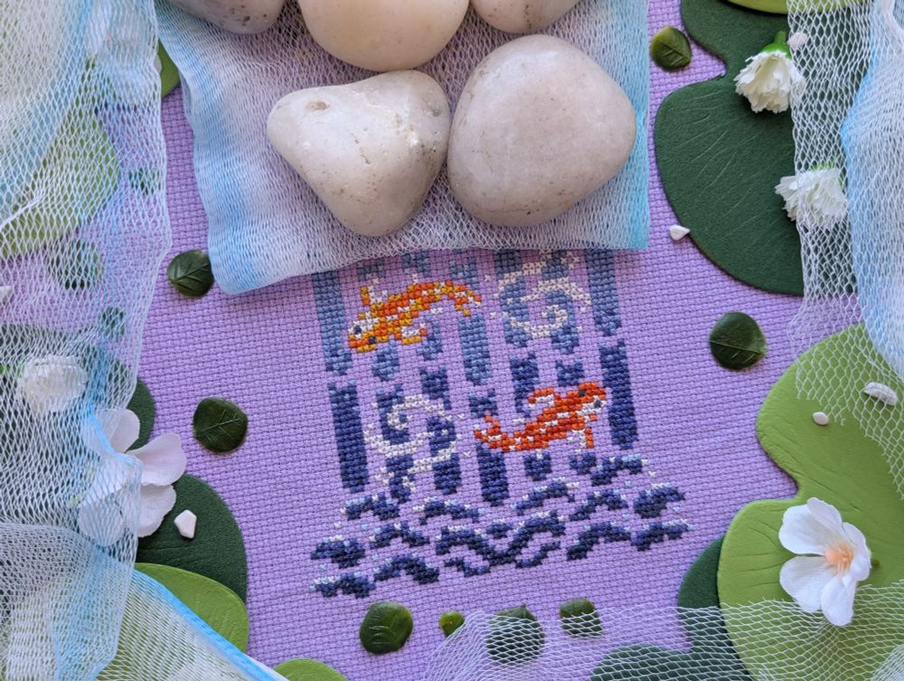 A partially covered cross stitch of a dark orange and light orange koi fish making their way up a waterfall on light purple aida. The cross stitch is surrounded by a blue and white mesh, lily pads, and white flowers.