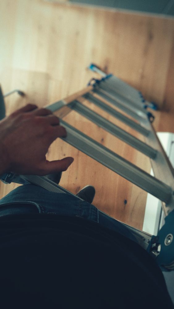 Foto von oben eine Leiter runter. Links meine Hand an einer Sprosse, ich lehne mich an die Leiter an.
Unten sieht man Holzboden und Kanten von Möbeln.