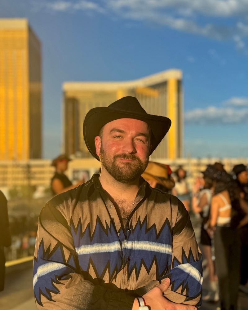 A man (me) standing in golden hour lighting at the Allegiant Stadium in Las Vegas with the Mandalay Bay Hotel in the distant background reflecting sunlight. He’s wearing a western shirt with spiky blue electric design and a black cowboy hat