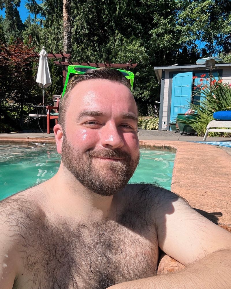 Photo of a bearded man with lots of body hair in the pool with a view of the pool surroundings in the background 