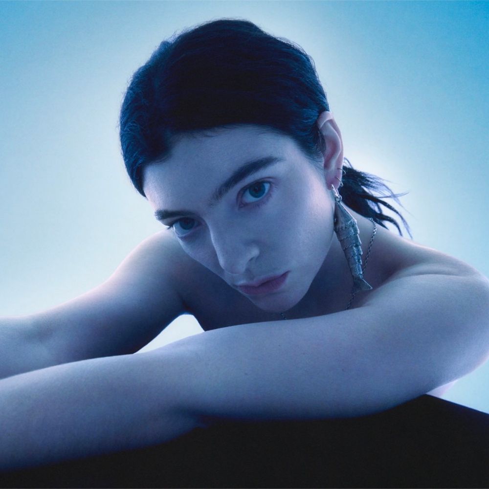 Lorde staring into the camera for a photoshoot for her new album. The color scheme is very blue and moody. She’s wearing a ponytail and some kind of dangly fish or mermaid tail earring. Werk