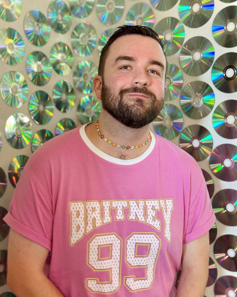 Me a bearded man standing in front of a wall covered in reflective CDs while wearing a Britney Spears jersey t-shirt and a flower choker because I’m a goddamn gay boy 