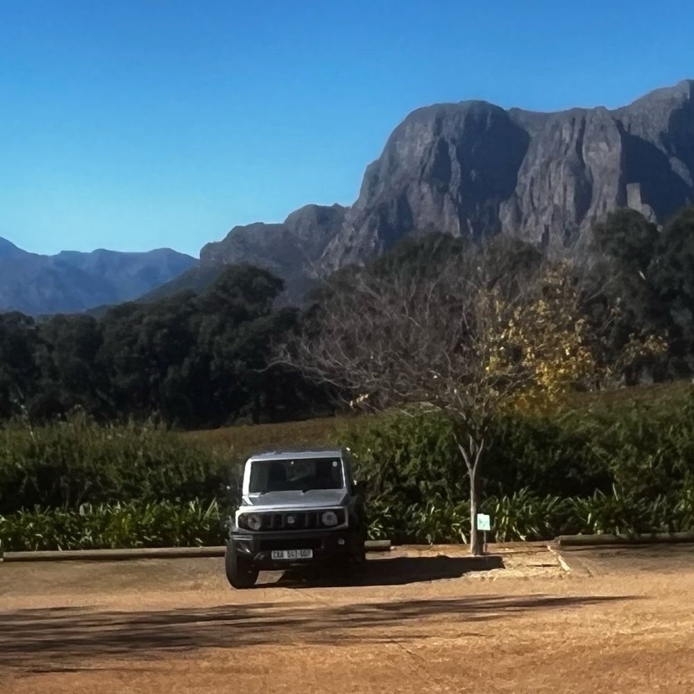 A small but perfectly formed 4x4 is parked with lush land and soaring mountains behind, all in blazing sunshine.