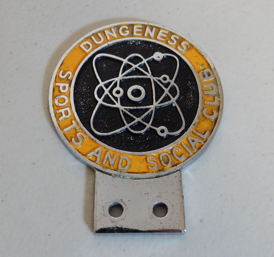 A vintage badge to be fixed to a car's grille. Around the symbol of an atom it reads 'Dungeness Sports and Social Club'