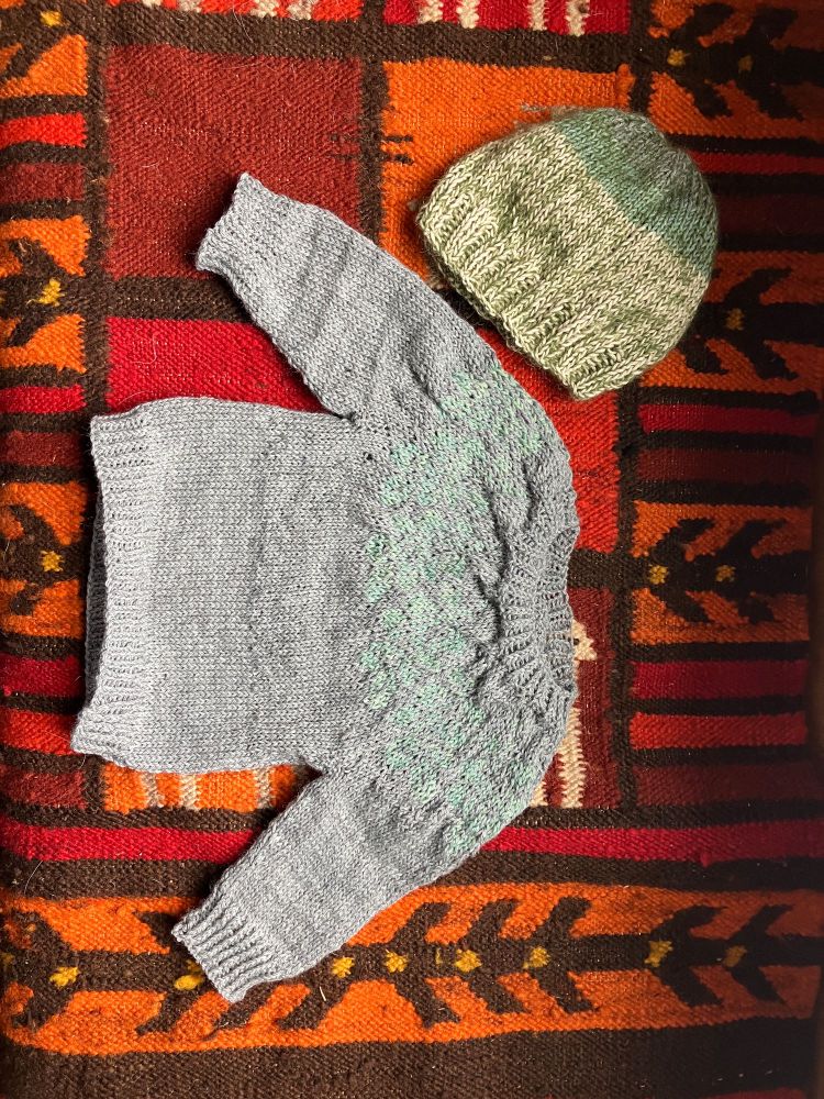 Hand knit baby clothes include a grey sweater and green beanie hat.