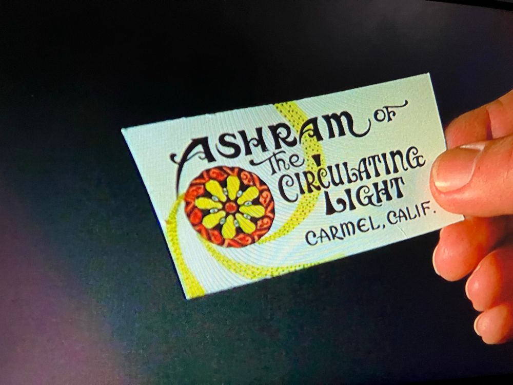 A psychedelic style business card held in a hand. The card reads “Ashram, the circulating light, Carmel, Calif”