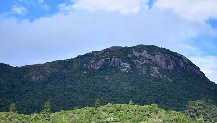 Foto de um Morro coberto de vegetação verde. Acima, um céu azul encoberto por nuvens. 