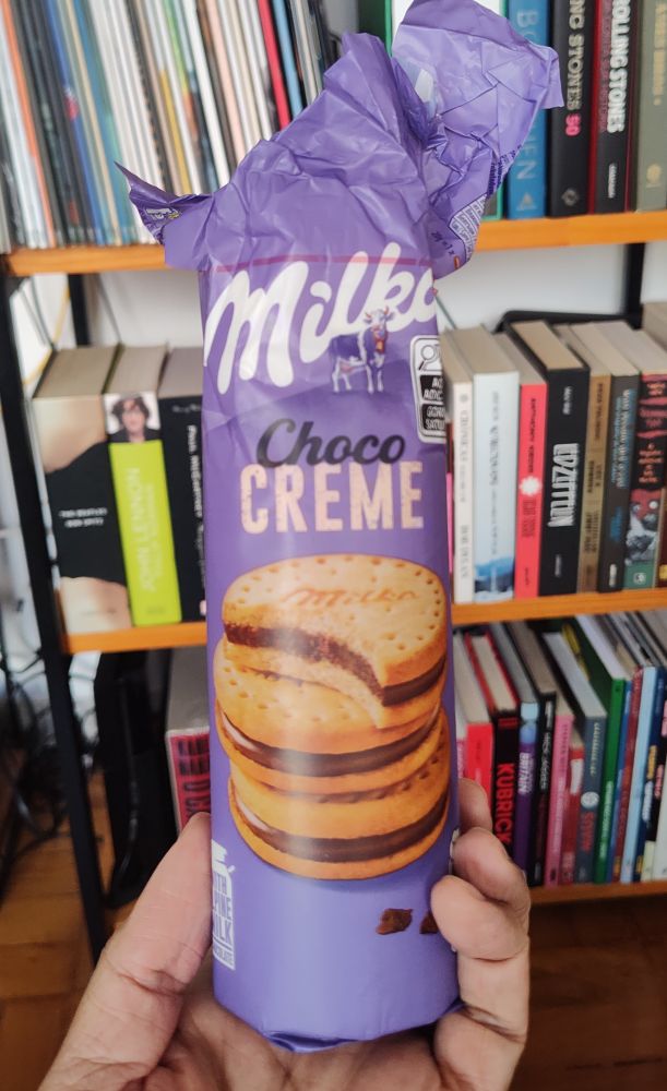 Minha mão segurando uma embalagem de biscoito recheado Milka. A embalagem é roxa com a foto de 3 biscoitos. 
