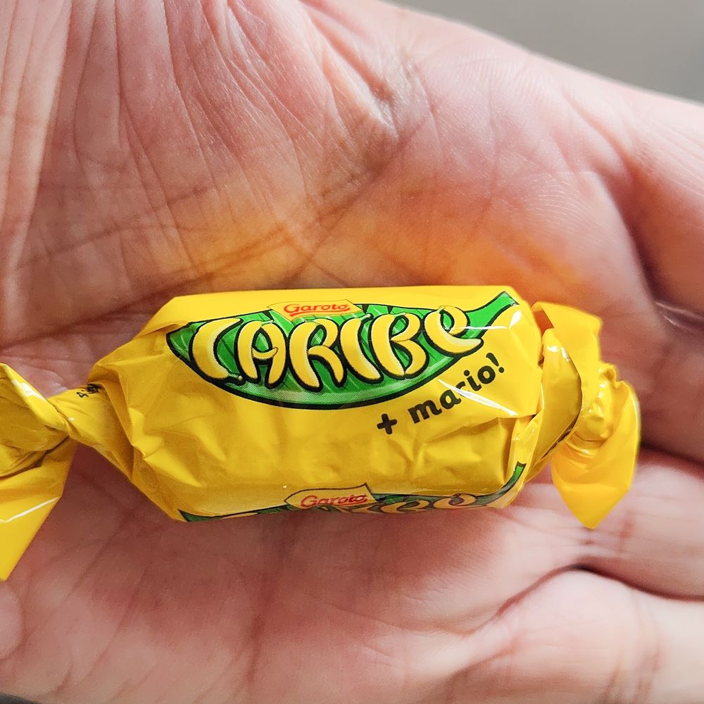 Uma mão aberta segurando um bombom Caribe. A embalagem é amarela com detalhes em verde.