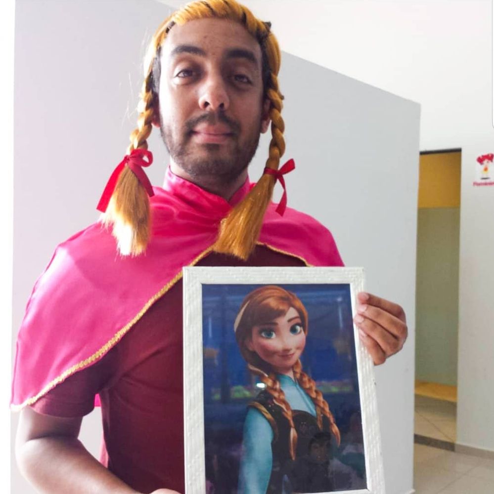 Foto minha fantasiado de personagem do filme "Frozen" , com peruca loira e uma capa rosa, segurando um quadro da personagem Anna do mesmo filme. 