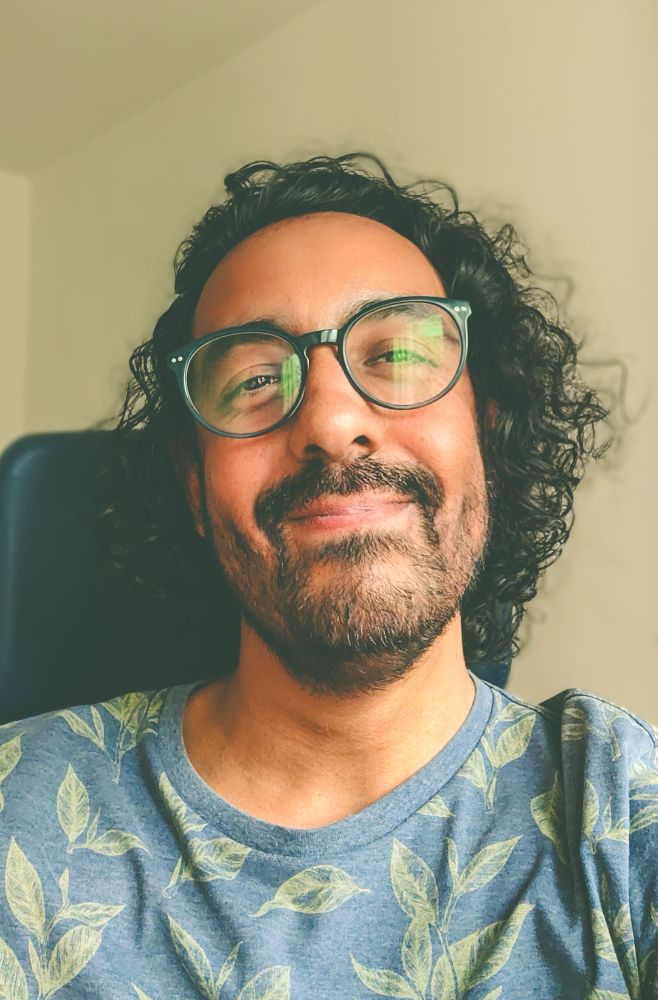 Foto minha com um leve sorriso de desespero. Homem negro com cabelos ondulados, usando óculos e uma camiseta azul com folhas verdes. 