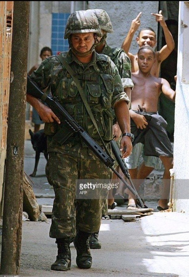 Homens do exército caminham en janto, ao fundo, vários garotos sem camisa fazem gestos obscenos em direção a eles. 