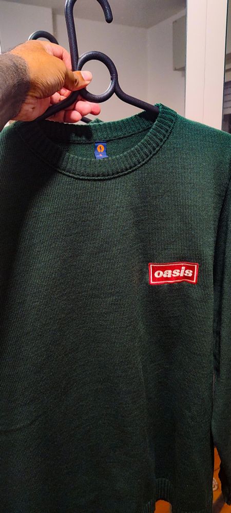 Minha mão segurando um suéter verde com o logo da banda Oasis em vermelho 