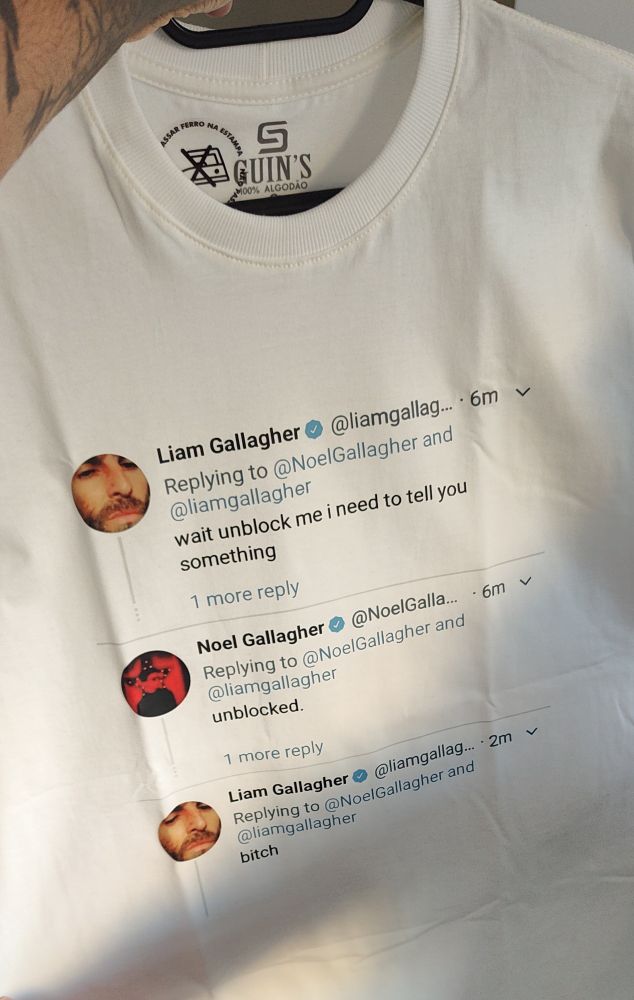 Alt text:
Foto de uma camiseta branca pendurada em um cabide. A estampa mostra uma sequência de tweets impressos, simulando uma conversa entre os irmãos Gallagher. No topo da camiseta aparece a etiqueta da marca “Guin’s 100% Algodão”. Abaixo, três tweets são exibidos: o primeiro tweet é do perfil de Liam Gallagher, onde ele pede para Noel Gallagher desbloqueá-lo porque precisa dizer algo; o segundo tweet é de Noel Gallagher, respondendo “unblocked.”; e o terceiro tweet volta a ser de Liam Gallagher, que responde com a palavra “bitch”.