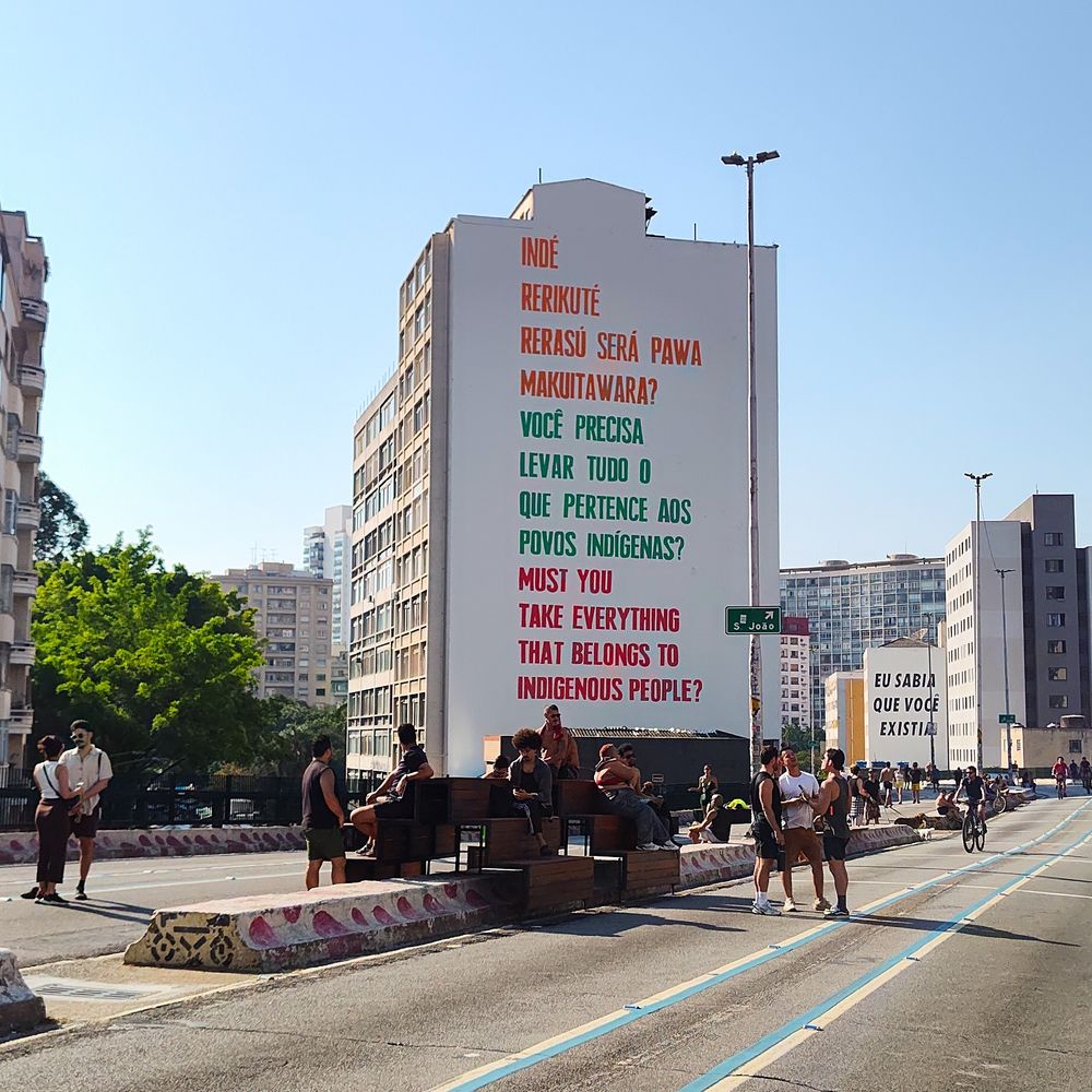 Um grande mural pintado na lateral de um edifício exibe uma mensagem em línguas indígenas, português e inglês. O texto diz:

“INDÉ
RERIKUTÉ
RERASÚ SERÁ PAWA
MAKUITAWARA?

VOCÊ PRECISA LEVAR TUDO O QUE PERTENCE AOS POVOS INDÍGENAS?

MUST YOU TAKE EVERYTHING THAT BELONGS TO INDIGENOUS PEOPLE?”

As palavras indígenas estão em laranja, a frase em inglês está em vermelho e o texto em português está em verde.

Na cena, pessoas caminham e conversam em um trecho do Minhocão, em São Paulo, que está fechado para carros e transformado em espaço de lazer. Algumas estão sentadas em bancos de madeira instalados sobre a via. Ao fundo, outro prédio exibe a frase “EU SABIA QUE VOCÊ EXISTIA”. O céu está limpo e azul, com árvores e vários prédios compondo a paisagem urbana.


