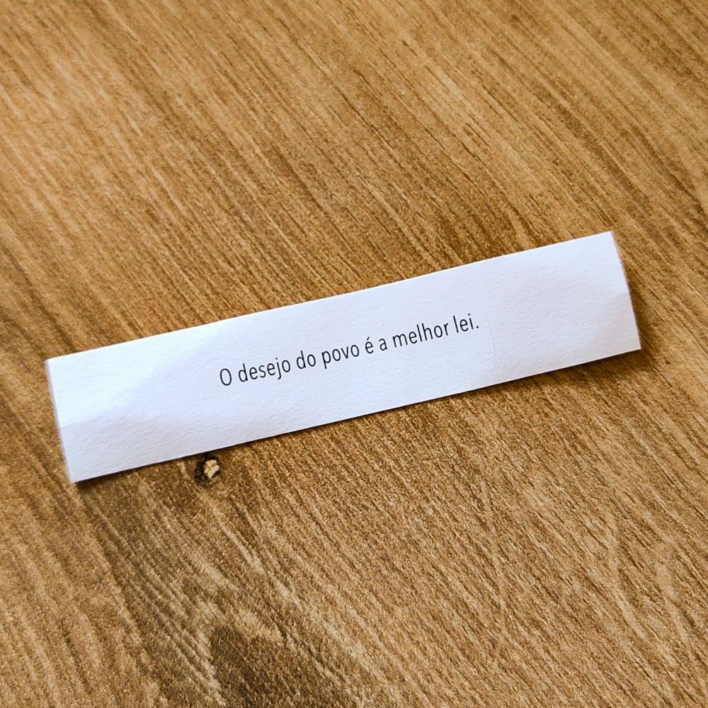 Papelzinho branco sobre uma mesa de madeira. No papel está escrita a frase "o desejo do povo é a melhor lei" em letras pretas. 