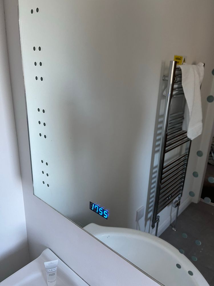 A picture of a mirror with  a digital clock embedded in it. The clock is half an hour slow.  