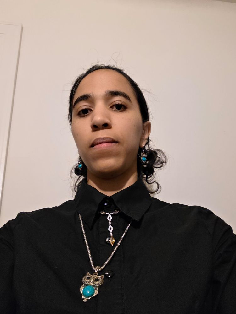 Selfie of Lex, a light-skinned person with a slim face, thick brows, and dark, curly hair. They are wearing a black, high-collared top and silver owl jewelry with turquoise accents. There is also a silvery choker with a pendant chain of three diamond-shaped pieces; one is topaz while the other two sparkle with tiny white gems. Probably rhinestones.

She tilts her head down at the camera, with a blank expression.