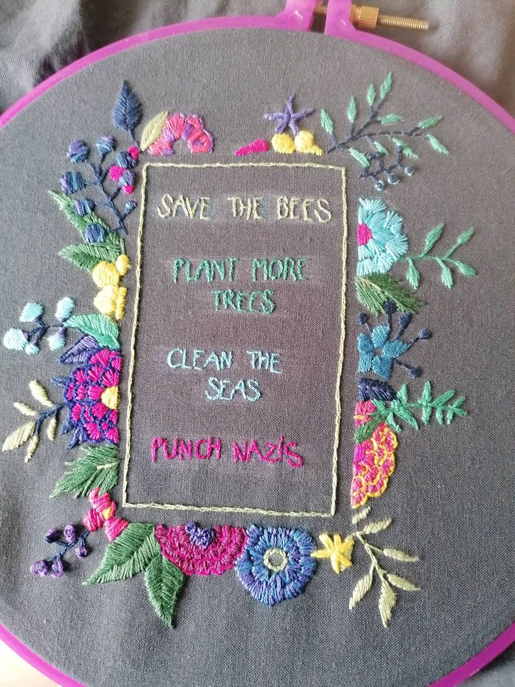 Broderie au fil coloré sur fond gris. Des fleurs et des feuilles encadrent un poème qui dit "save the bees, plant more trees, clean the seas, punch nazis" (en français : sauvez les abeilles, plantez plus d'arbres, nettoyez les mers, frappez les nazis). 