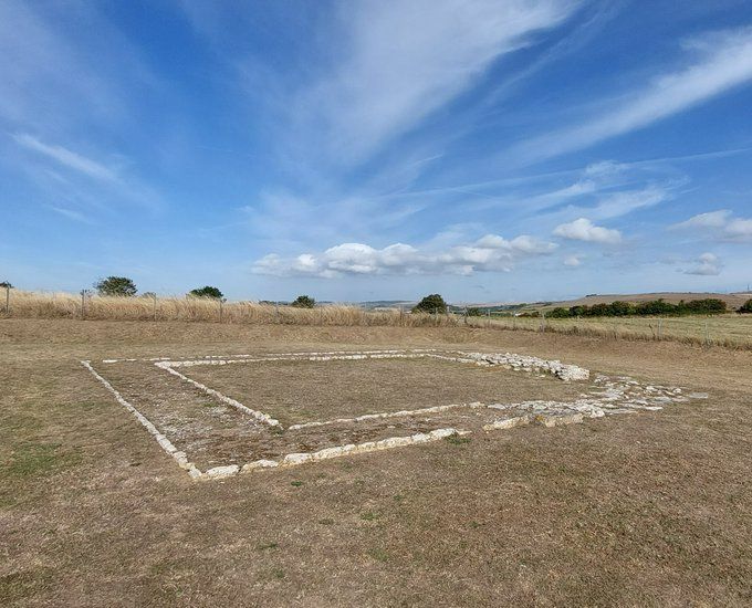 Photo of a ruined Roman temple, visible in the form of two concentric squares of white stone on grass, beneath blue skies