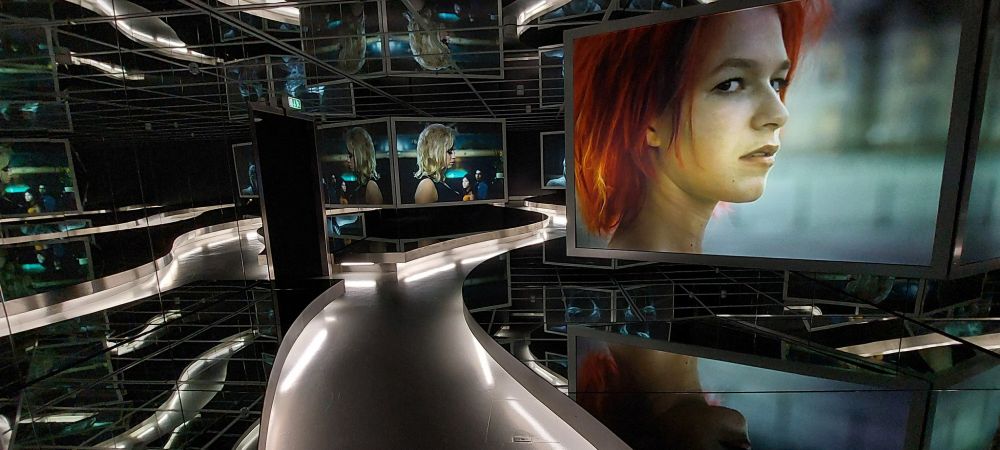 Photograph of one of the galleries inside the Deutsche Kinemathek, Berlin, showing multiple screens and mirrors around an illuminated walkway, with a scene from the film 'Run, Lola, Run' showing red-haired actress Franka Potente