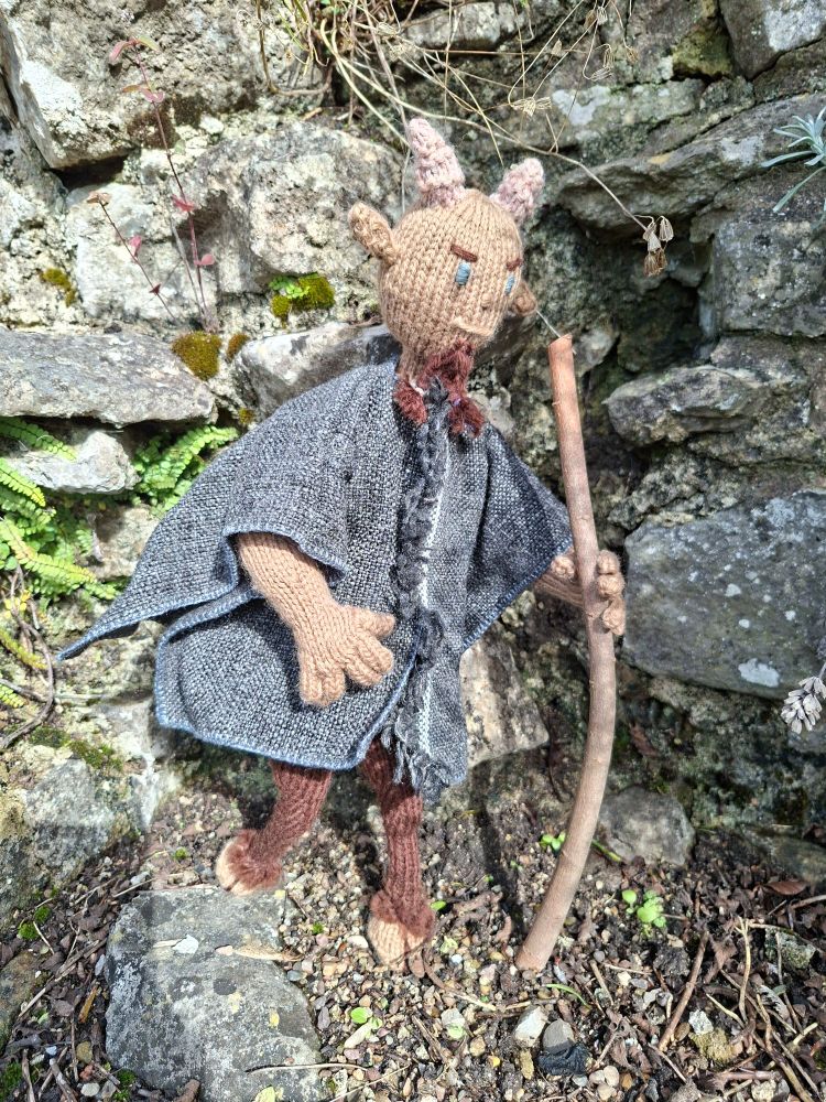 A knitted figure of a faun taking a walk in a rocky garden. He's dressed in a cloak and holding a walking stick