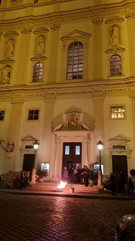 The Jesuit / University Church of Viena. Proof that science and religion can go hand in hand.

It is after sun-set. Lantern stand in front of the brilliant white church fassade. It is a high baroque church. So even the white outside has alcoves with several life-sized and very detailed statue.

In front of the church is the tiniest easter fire I have ever seen. A fire place the size of a bigger dinnerplate with a few logs. The fire illuminates the cobblestone of the piazze in front of the church. About 8 people are standing around the scenery.

It's actually a pretty scene tinged with soft yellow.