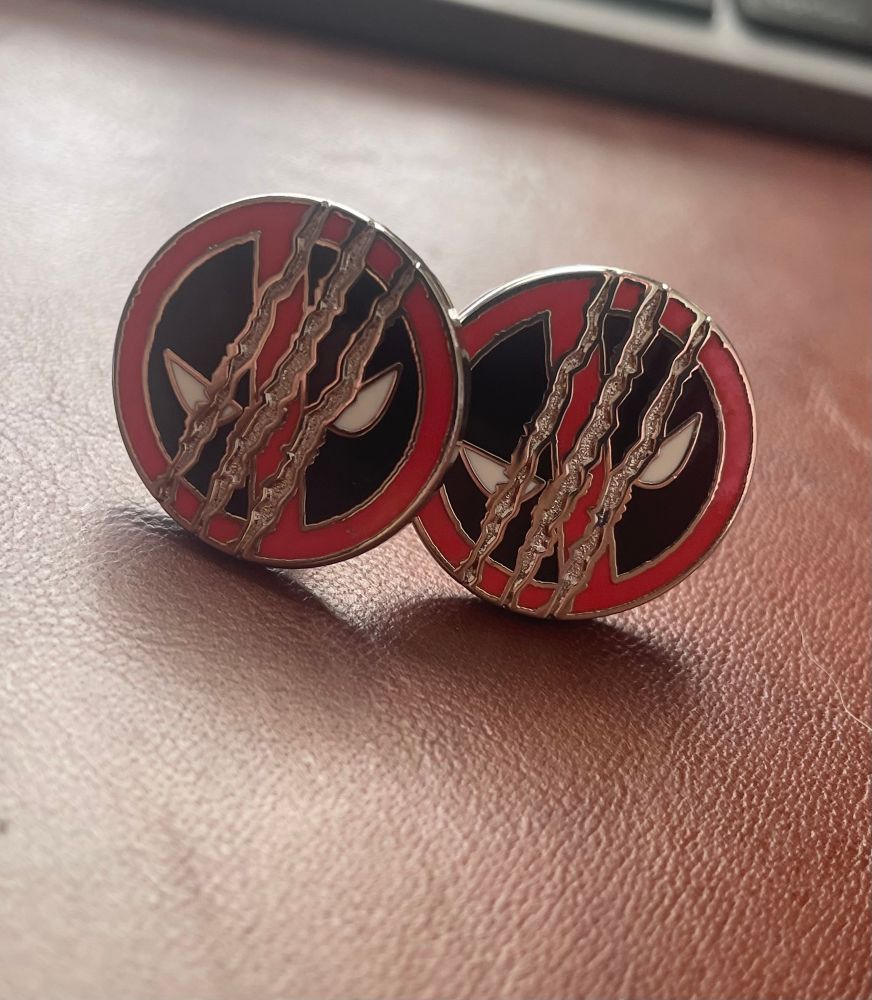 a picture of two cufflinks with the Deadpool logo on their faces, a red mask background with two eye eyes surrounded by black overlayed with three claw marks through the front of the masks, meant to symbolize Wolverine.