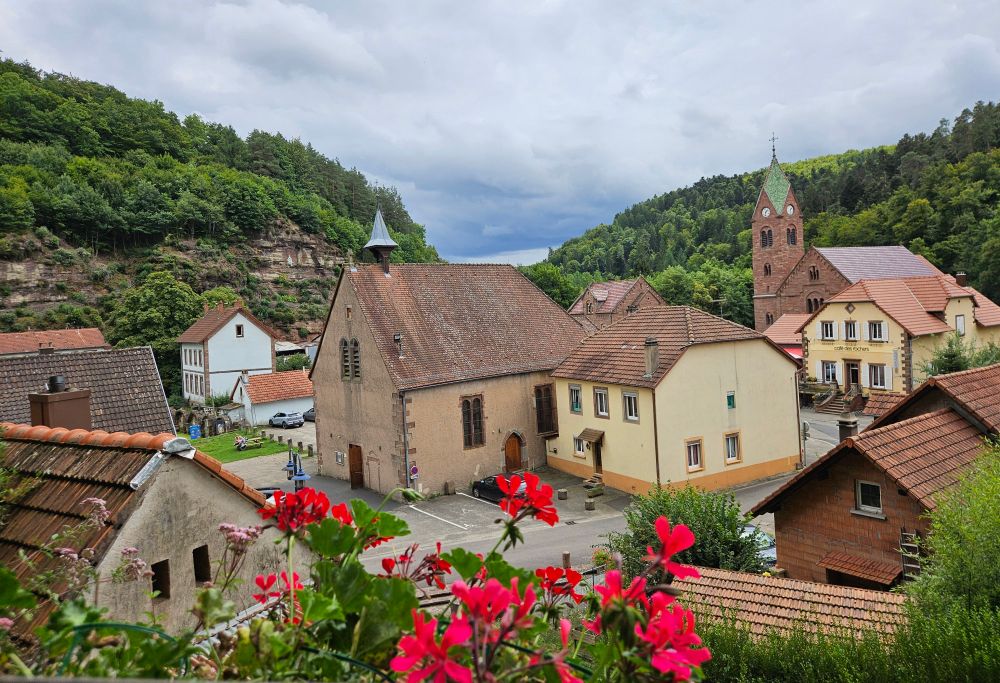 View of the village of Graufthal: the old Protestant church in the front, the more recent Catholic church in the back, hills with woodland and steep rocks in the background, blossoming geraniums in the front 