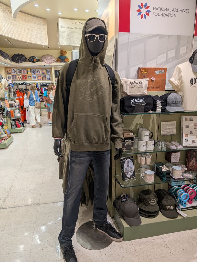 Photograph of a mannequin in a gift shop. It is wearing a hoodie labeled Record division and a hat with the same. Above it and to the right is below Bell of the National Archives foundation. Beside, it is an assortment of hats, shot glasses, mugs, socks, stickers, mugs, kitchen magnets, and coasters with the same insignia.