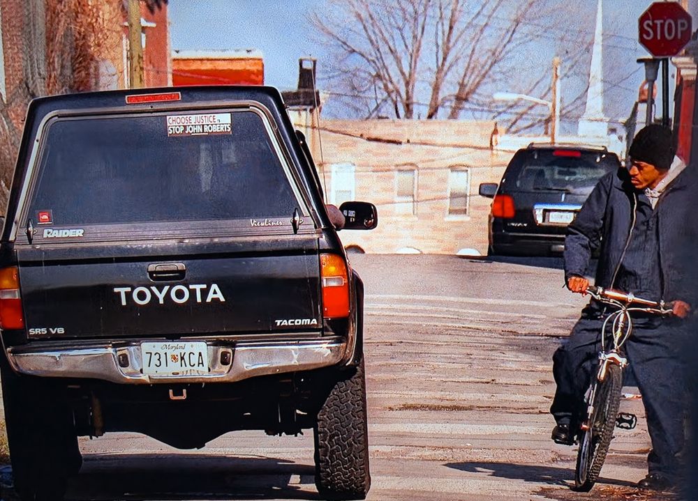 screenshot from the wire, season 4, episode 8.

Outdoor scene - on a street. On the right is a man on a bike.

On the left is a pickup truck with a camper top. On the back glass is a bumper sticker that reads "Choose Justice  / Stop John Roberts"