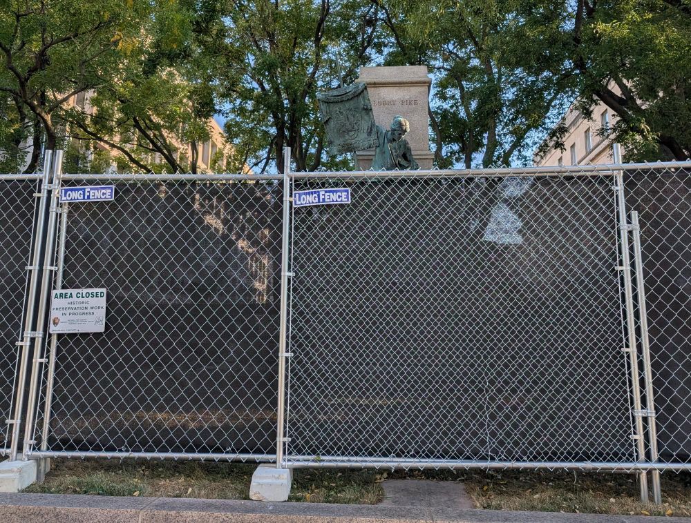 Photograph of fencing around the base of the Albert Pike memorial in DC, which is visible above the fencing. A sign on the fence bears NPS logo and reads 