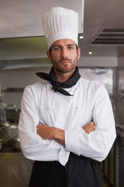 picture of a serious-looking chef looking at the camera, wearing a toque and standing in a stainless steel commercial kitchen