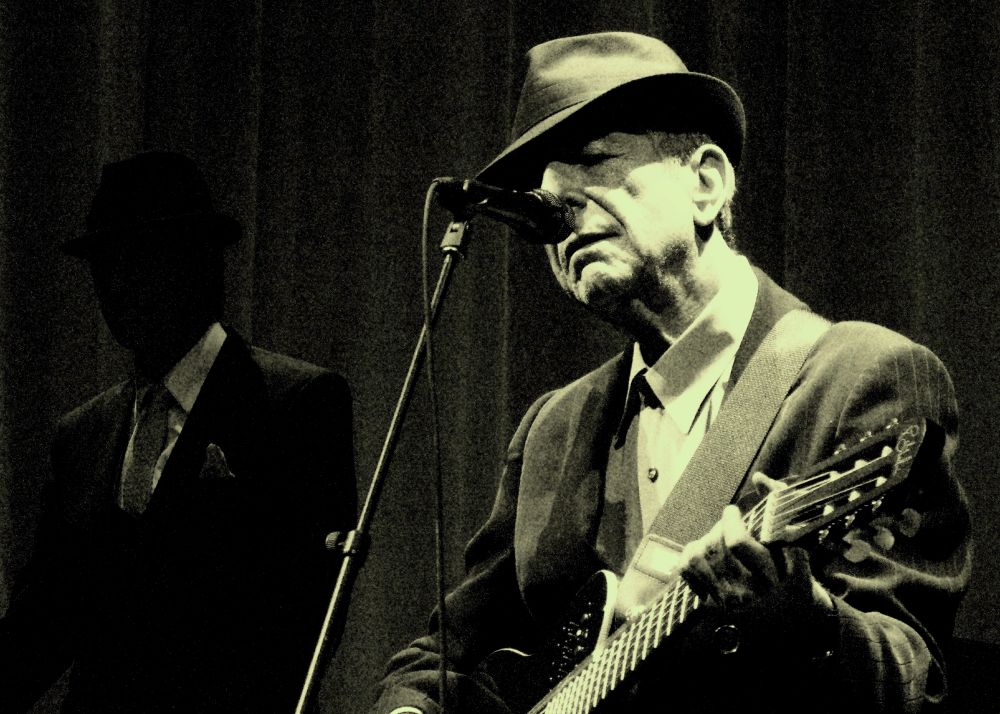 Singer Leonard Cohen, eyes closed, in front of mic, fedora in place, playing acoustic guitar.