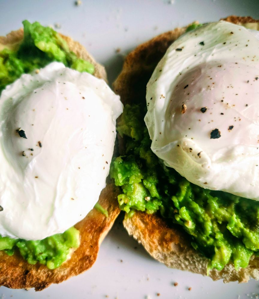 Close-up photo of two toasted English muffin halves topped with bright green mashed avocado and soft poached eggs. The eggs have smooth white surfaces with a few specks of black pepper. The muffins are golden brown around the edges, and a few crumbs and pepper flakes are scattered on the plate.