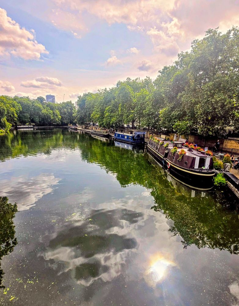 Little Venice in London, showing a tranquil canal under a partly cloudy sky, with a fiery, glowing ball of sunlight reflected on the water’s surface. The calm canal mirrors the sky and surrounding greenery, with lush trees lining both banks. On the right, several moored narrowboats, decorated with colourful flowers on their roofs, rest beside a brick wall. The left bank is shaded and natural, while in the distance a modern high-rise building peeks above the treeline, adding an urban touch to this peaceful waterside scene.