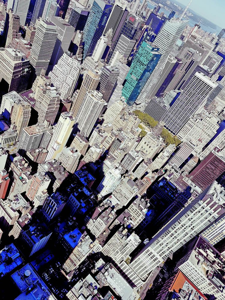 Aerial view of Midtown Manhattan filled with densely packed skyscrapers casting long shadows across the city streets. The dark triangular shadow of the Empire State Building stretches prominently across the surrounding buildings, contrasting with the sunlit rooftops. The photo captures a mix of modern glass towers and older stone high-rises, with the Hudson River faintly visible in the background under a clear sky.