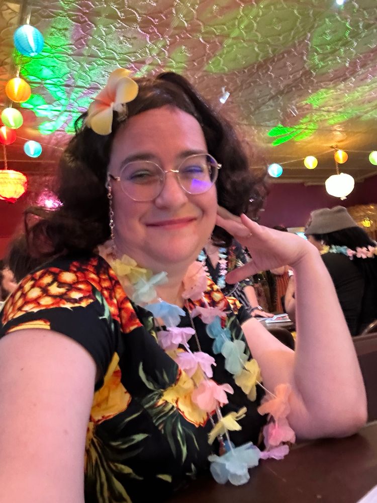 Middle aged woman with curly hair dressed in a black Hawaiian dress with pineapples, a yellow tropical flower in her hair, a lei of multicolored flowers, and wire framed glasses on a gold chain. She is smiling at the camera with her left hand on her left cheek. Her look is early 1960’s. She is sitting in a large room decorated for a luau.