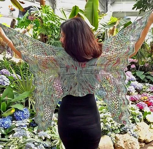 A dark haired woman with her back to us, holds out the points of a shawl shaped like a butterfly. She stands in a flower garden.