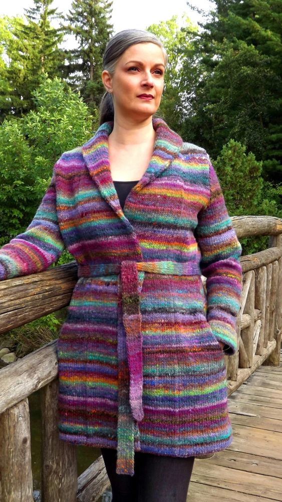 A woman wears a striped, self-belted knitted coat with lapels, and pockets. The colours are mostly bright purple and greens. She stands on a log bridge.