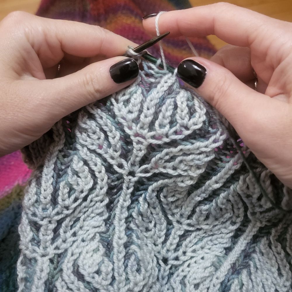 Hands knitting a two colour fabric of light gray and medium grayish-teal blue. It is an unfinished hat in brioche stitch.