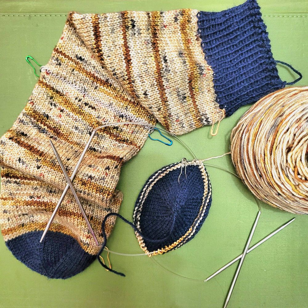 In progress socks of brown MC and navy CC against a green background. One sock only needs the heel to be worked. The second sock has only the toe complete