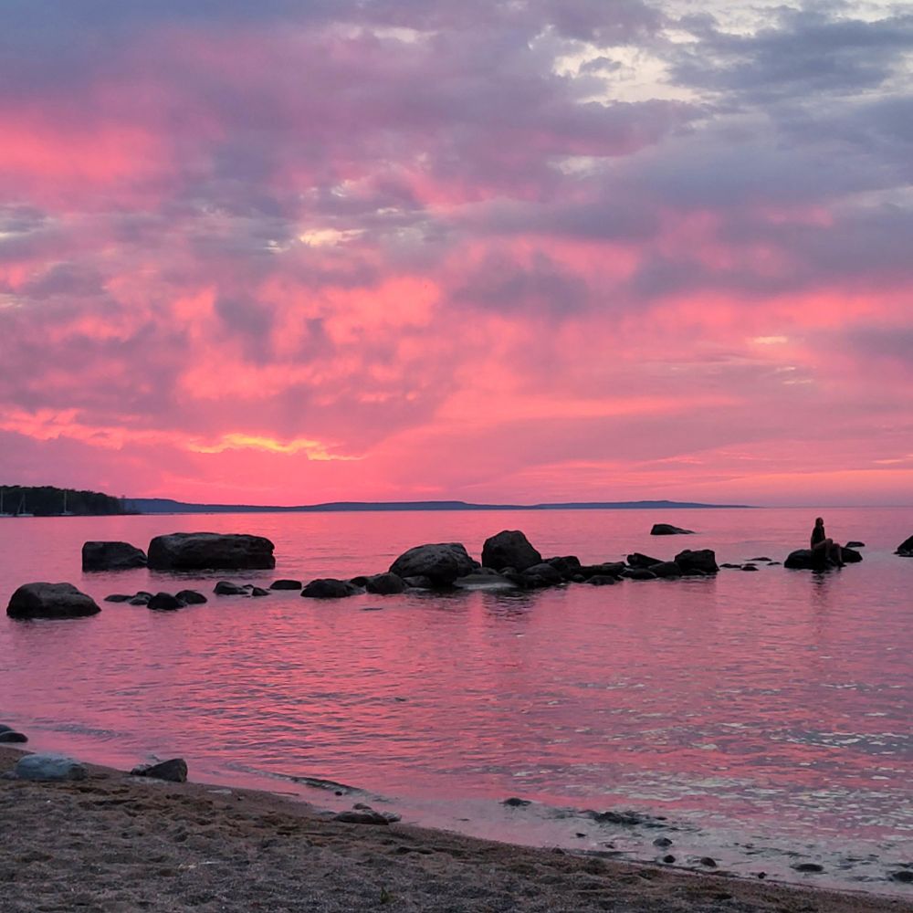 A deep pink sunset with light purple clouds over the water at a beach. There are large boulders in the water and a small figure of a person sit on a far boulder.