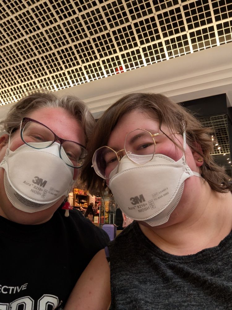 A European-American woman and her non-binary partner smile while wearing masks in a mall setting.
