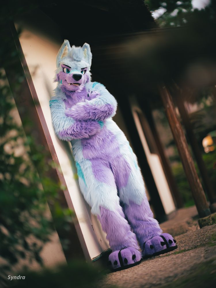 Keenora leaning at a wall, in his Japanese styled fursuit in a Japanese styled setting. He has his tongue out a bit bit a very sneaky expression and crossed arms.