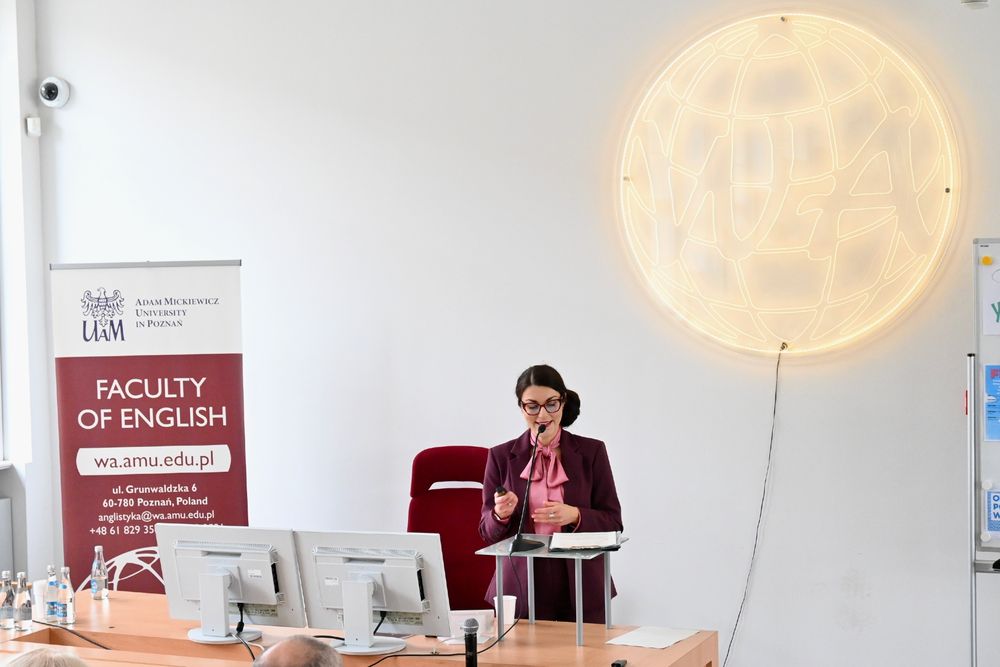 A woman in a violet jacket and a pink shirt with a brown bun delivering a lecture. Behind her a light illumination saying "WA". On the left a banner saying "Faculty of English".